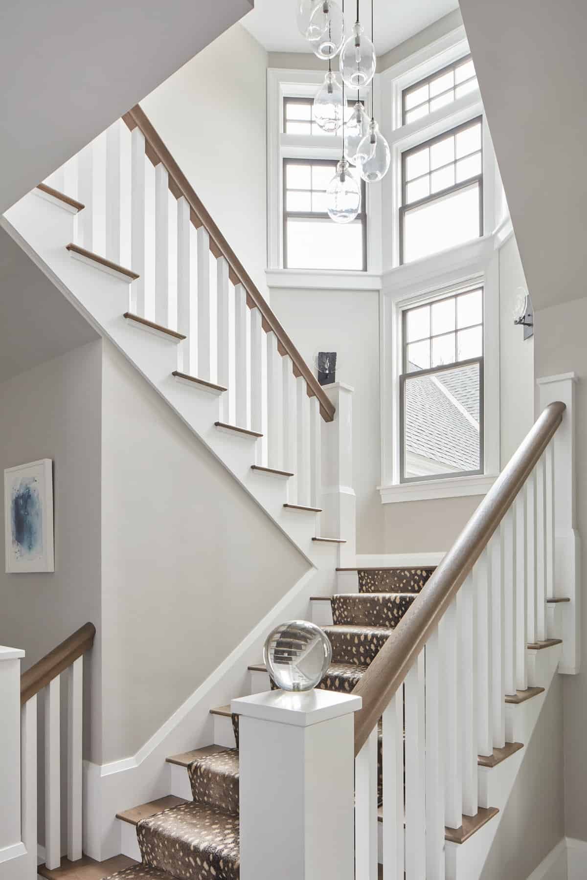 Interior view of a bright staircase with white walls, wooden banister, and windows letting in daylight. Modern hanging glass light fixtures are visible.