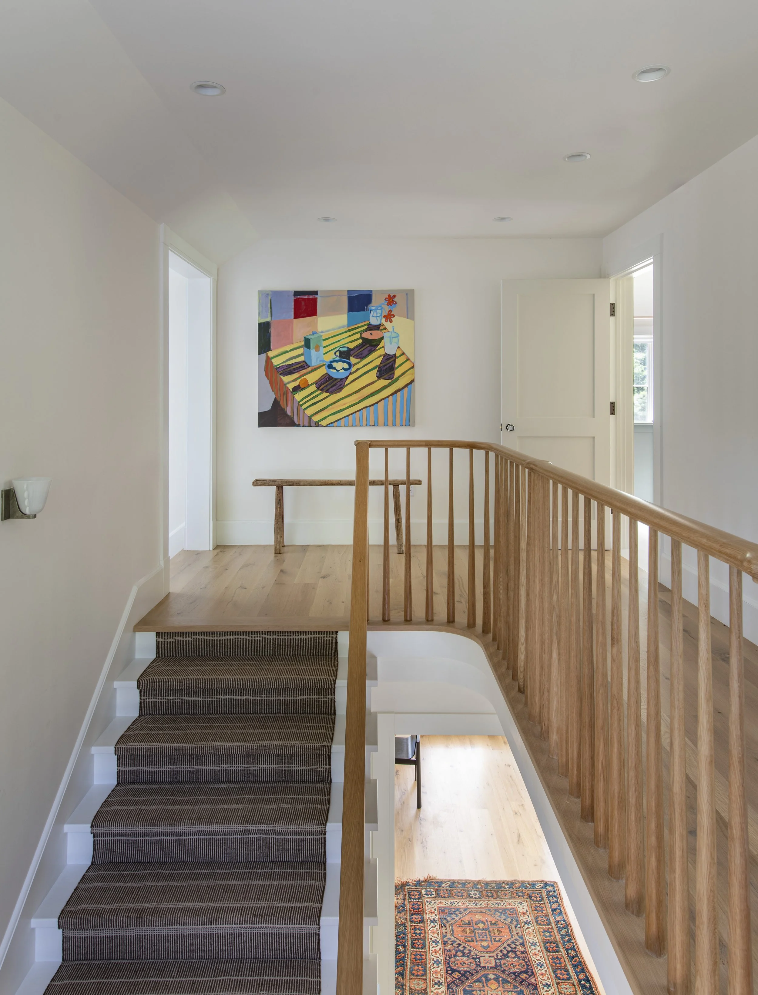 Interior view of a modern staircase landing with a wooden railing, a colorful abstract painting on the wall, a small wooden bench, and a vibrant patterned rug on the hallway floor, natural light coming through a window.