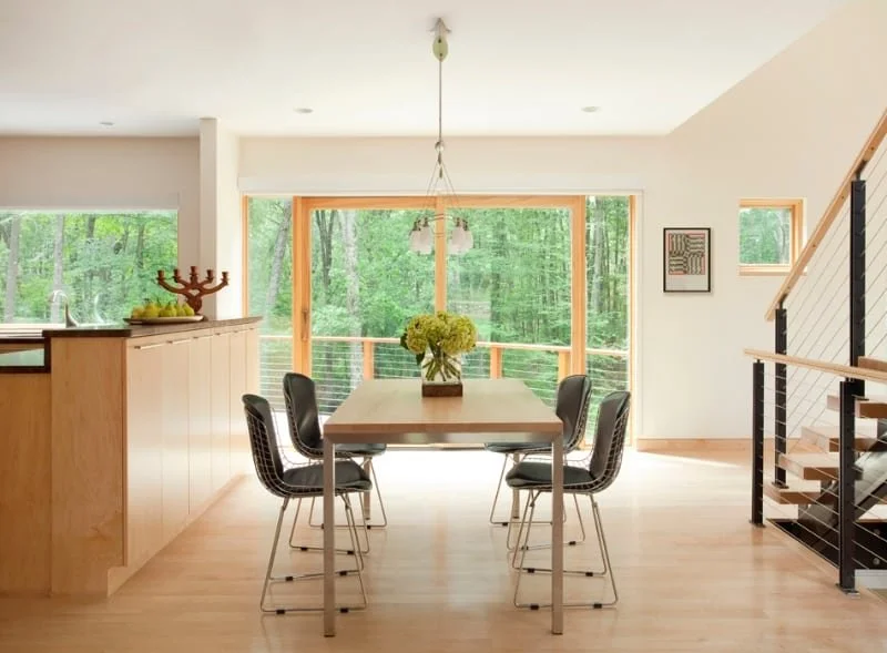 Modern dining room with wooden table, six black chairs, large window overlooking a green forest, and a staircase to the right.