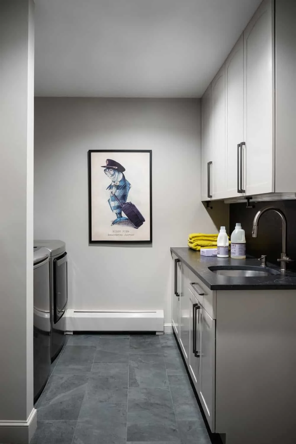 A laundry room with white cabinets, a black countertop, a sink, two bottles of cleaning supplies, yellow towels, and a framed art print of a person in a black hat with a suitcase.