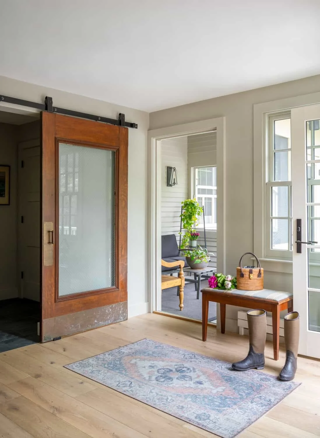 A cozy entryway with a sliding barn door, a rug, a small table with a basket and flowers, and boots on the floor near the door. A sunroom with plants and a chair is visible through the open door.