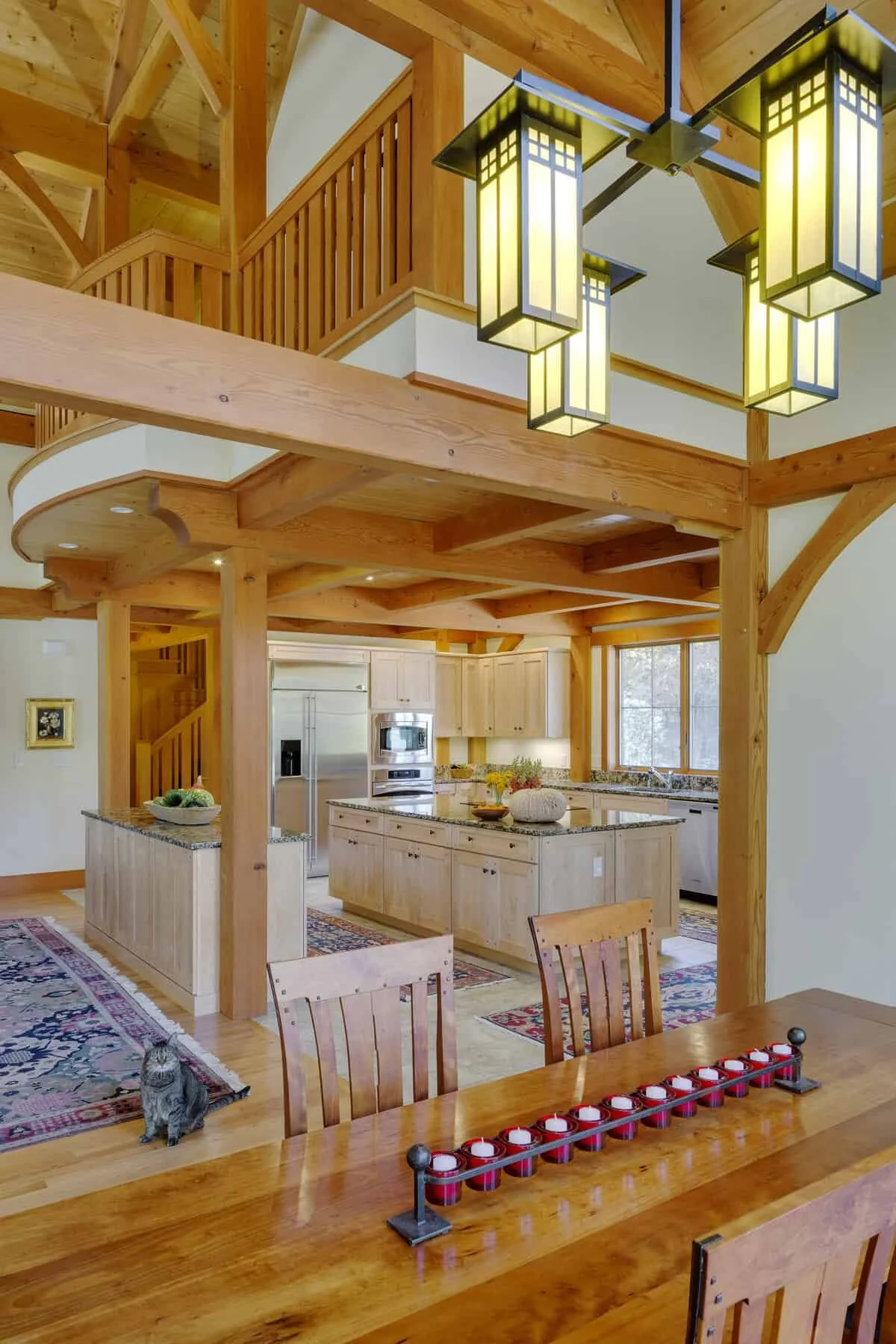 Interior of a wooden home with a kitchen and dining area, featuring exposed wooden beams, a kitchen island with a granite countertop, and a hanging modern chandelier. A gray tabby cat is sitting on a colorful rug near a wooden dining table with red t