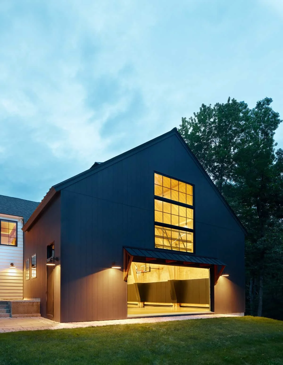 A modern black garage with large window displaying an illuminated indoor basketball court, set against a backdrop of trees and a cloudy sky.