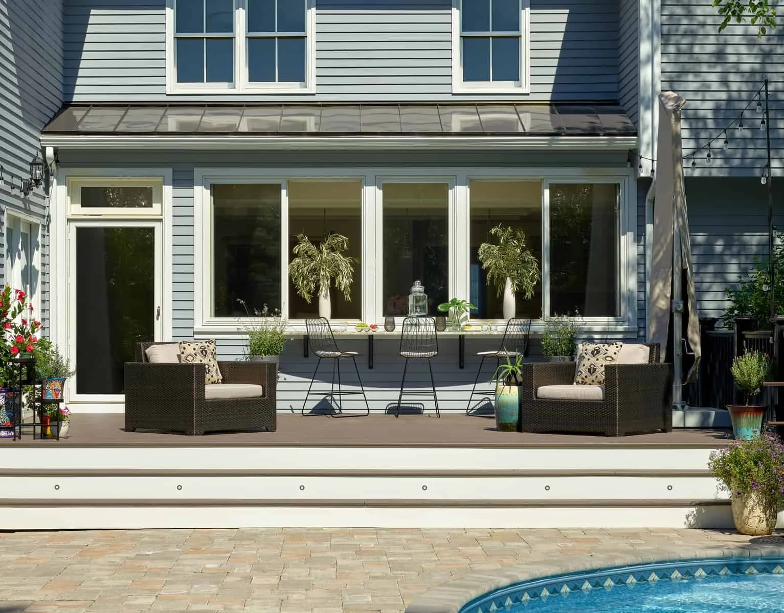 Backyard patio with outdoor seating, barstools at a high table, potted plants, and a swimming pool in the foreground.