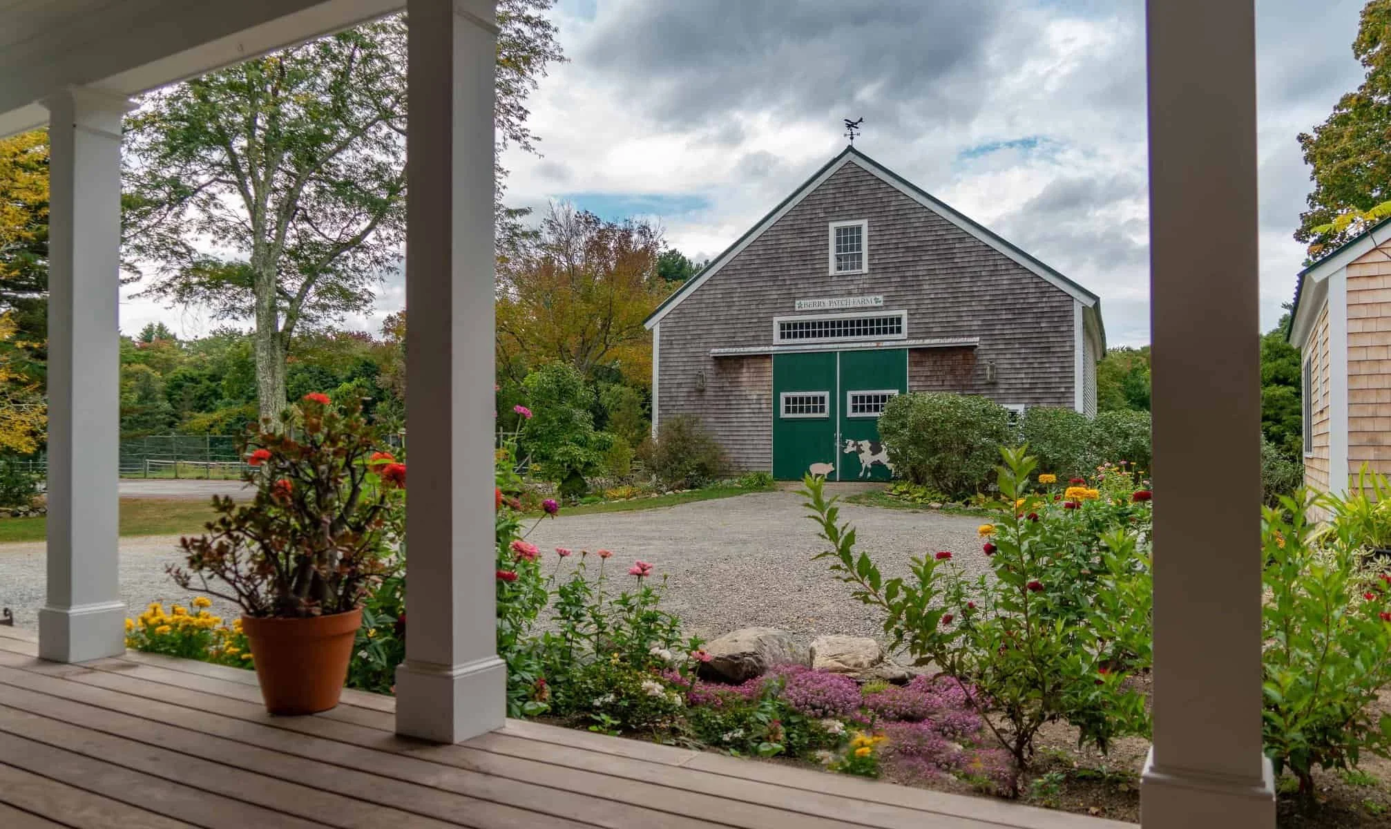 View from a porch with white columns overlooking a garden with potted flowers, a gravel path, and a large barn with green doors and a weather vane on top, surrounded by trees with fall foliage and cloudy sky.