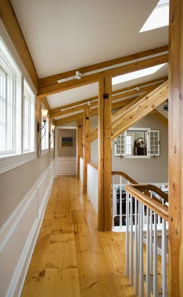 Looking down a hallway with wooden beams, hardwood flooring, and windows on the left side, featuring a staircase with a white railing and a ceiling skylight.