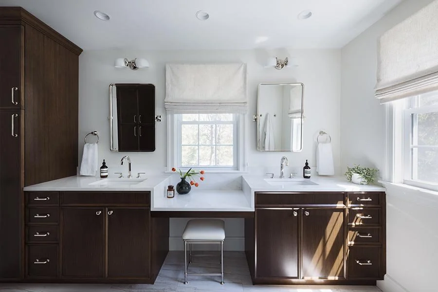 A bathroom with dark wood cabinets, white countertops, two sinks, two mirrors, and a window between the sinks with a small white chair underneath. There are two small wall-mounted lights above the sinks, and a few decorative items such as a small pla