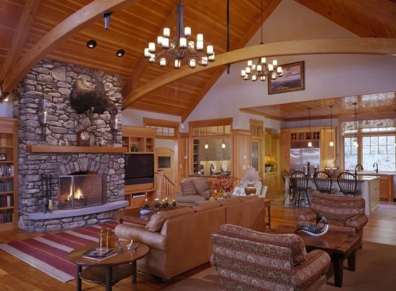 A cozy living room with wooden vaulted ceilings, a stone fireplace with a mounted animal head, and a chandelier. The space includes a sofa, armchairs, a TV, and a kitchen area with barstools.