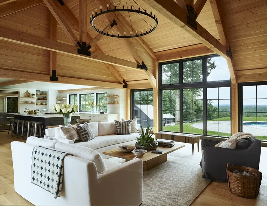 Living room with high wooden ceilings, large windows, and modern furniture, including white sofas and a dark armchair, with a view of an outdoor pool and greenery.