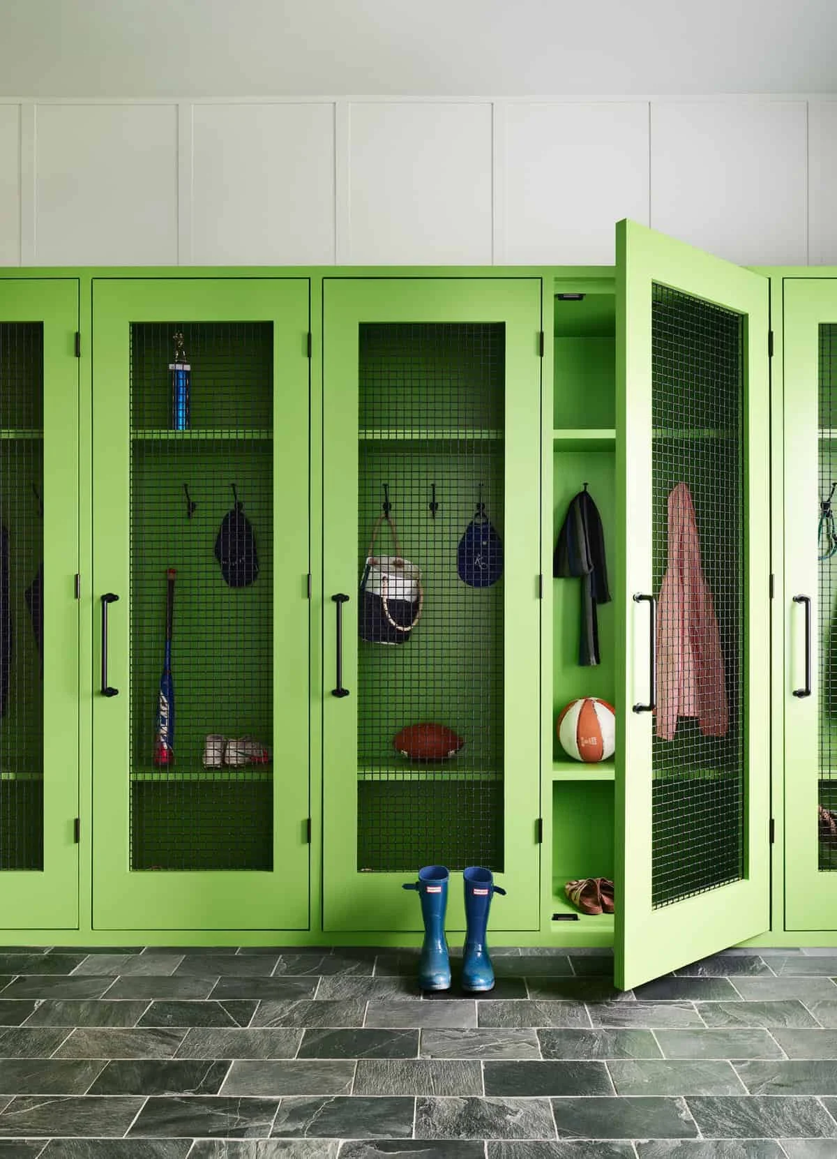 Green lockers with black mesh doors in a sports locker room. Items such as a baseball bat, gloves, soccer ball, rugby ball, basketball, and boots are stored inside. Blue rubber boots are placed on the floor in front of the lockers.