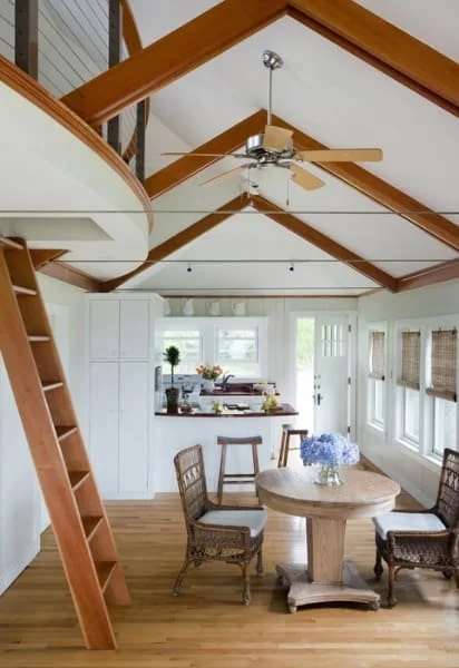 Open-concept living space with white walls, wooden beams on the ceiling, windows with blinds, a dining table with four chairs, a ceiling fan, and a small kitchen area in the background.