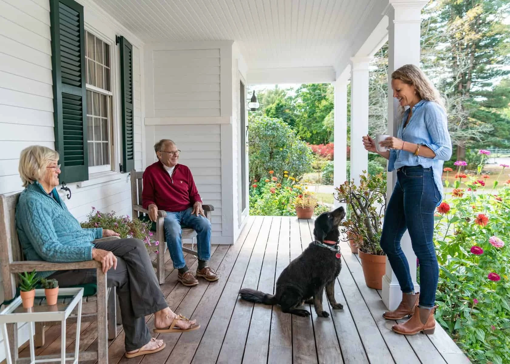 Three older adults sitting on porch with woman standing with dog and coffee mug, surrounded by plants and flowers, in daytime.