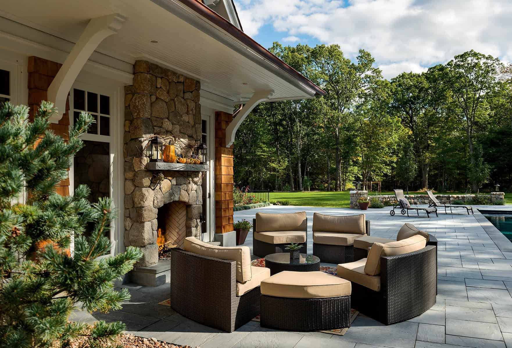 Outdoor patio area with a sitting area, stone fireplace, and a swimming pool with lounge chairs, surrounded by a grassy yard and trees under a partly cloudy sky.