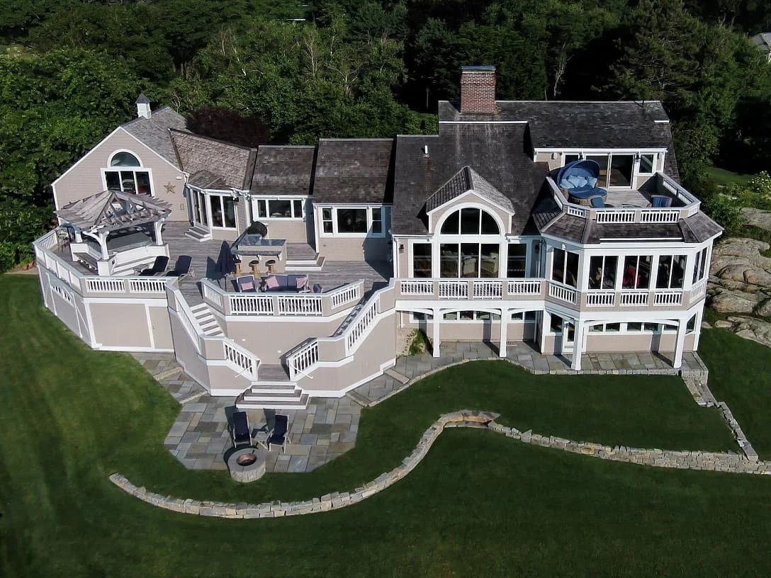 A large, multi-level house with a spacious wooden deck and an outdoor firepit area, surrounded by lush green lawn and trees.