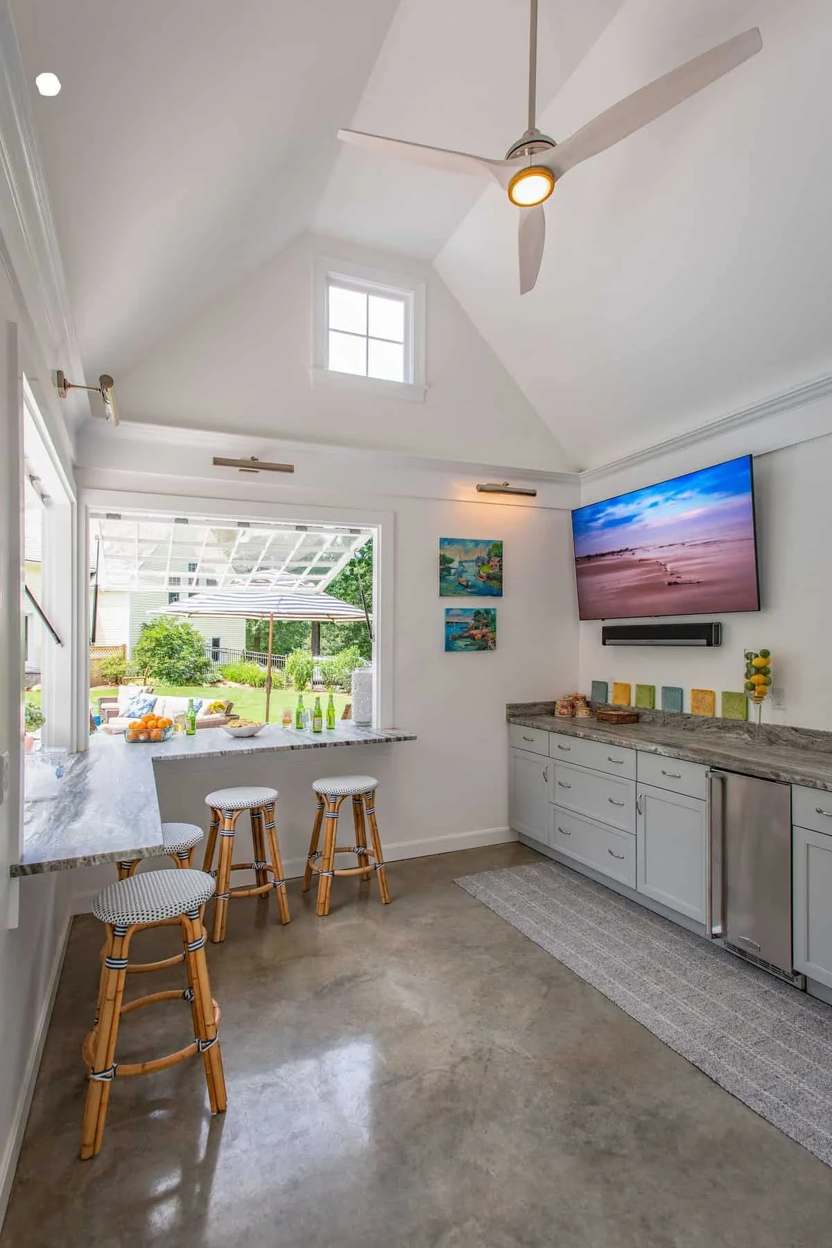 Modern kitchen with white cabinets, marble countertops, and a large window overlooking a backyard with an umbrella, fruit, and beverages. A wall-mounted TV displays a beach scene, and there are colorful artwork pieces above the counter. Pendant light