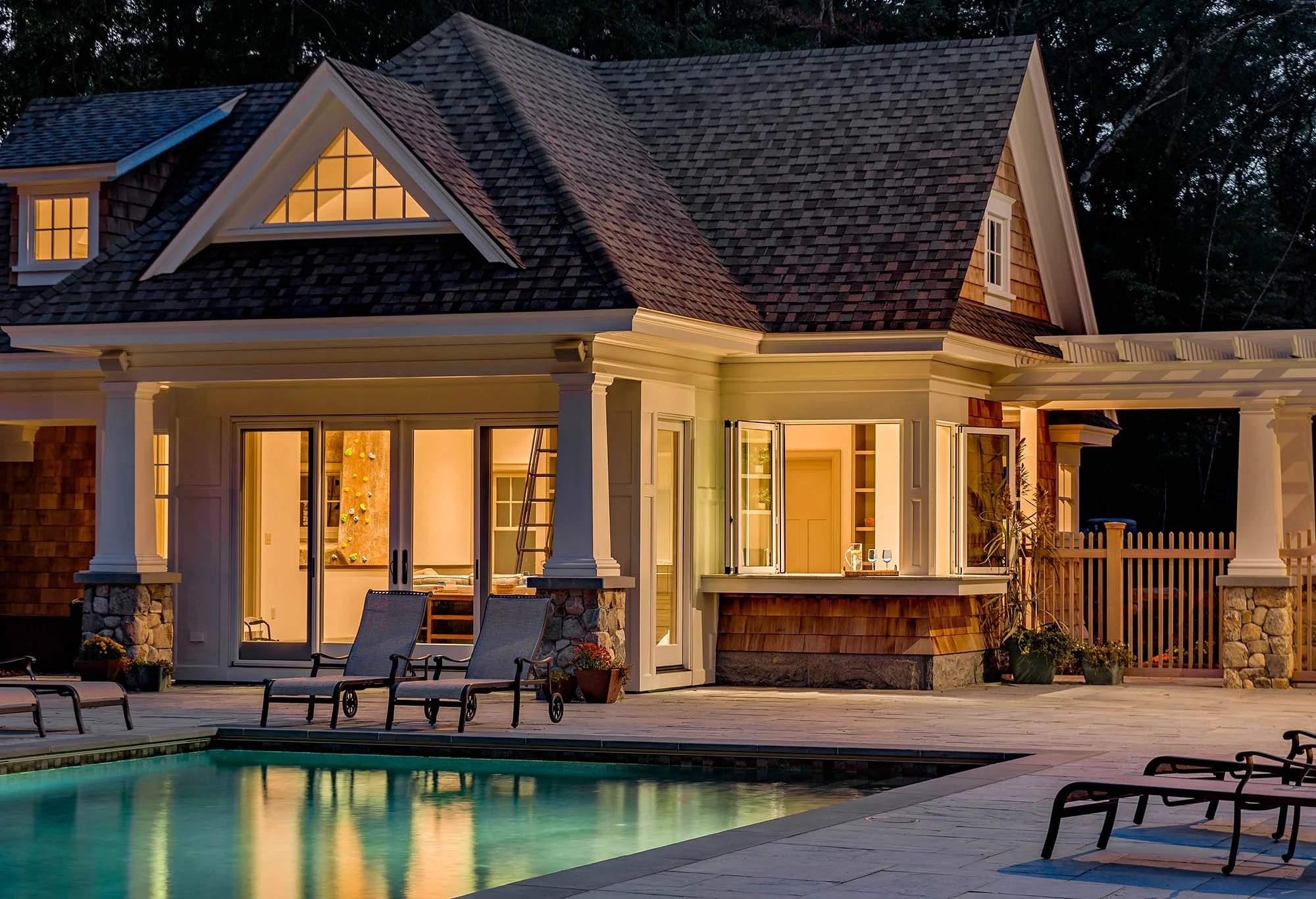 A house with a shingled roof and large windows illuminated from inside, featuring a backyard with a swimming pool and lounge chairs during dusk or evening.
