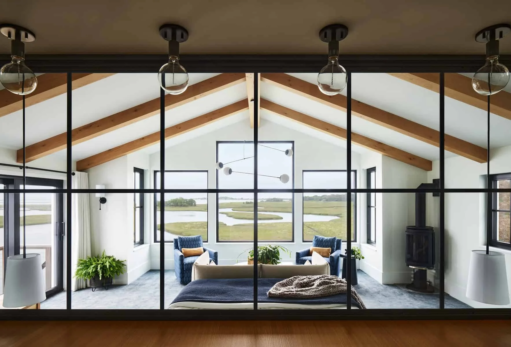 Living room with large windows and a view of a river and landscape, featuring a sofa, blue armchairs, modern chandelier, and fireplace.