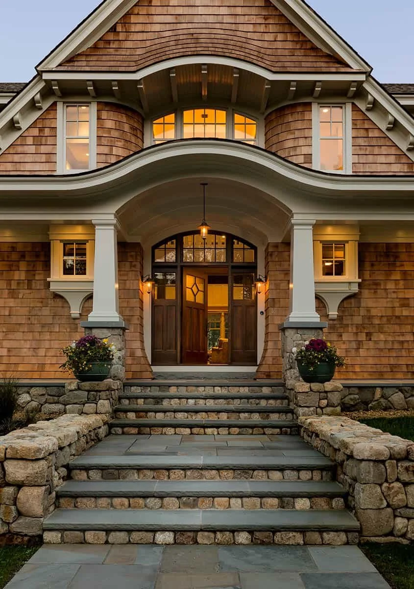 Front entrance of a large house with stone steps, wooden siding, white columns, and lit windows, taken at dusk.