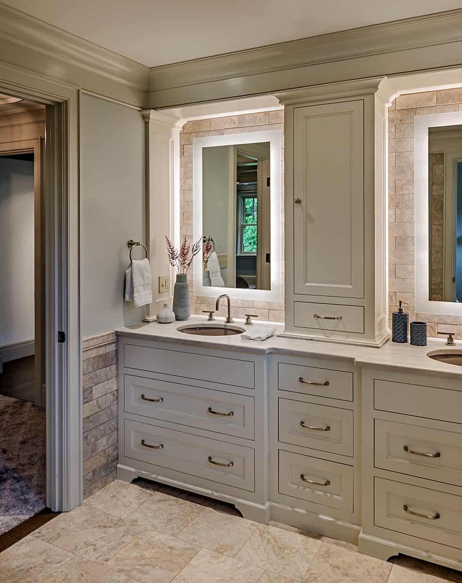 A double bathroom vanity with beige cabinetry, dual mirrors with backlighting, a tan stone backsplash, and decorative vases with dried flowers. To the left, a towel hangs on a ring next to electrical outlets. Part of a doorway leads to a room with wo