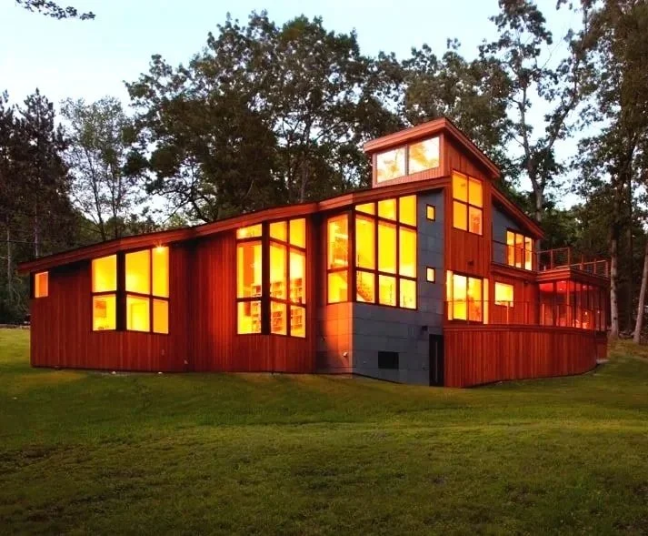 Modern contemporary custom home with a combination of wooden and gray exterior walls, multiple large windows illuminated from inside, set on a green lawn with trees in the background.