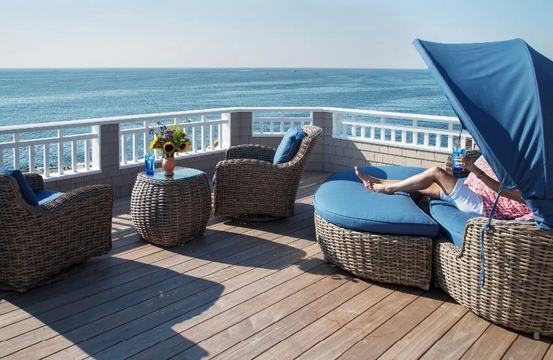 A seaside view from a wooden deck with wicker outdoor furniture, including two armchairs, a side table with a vase of flowers, and a lounge chair with a blue canopy, overlooking the ocean.