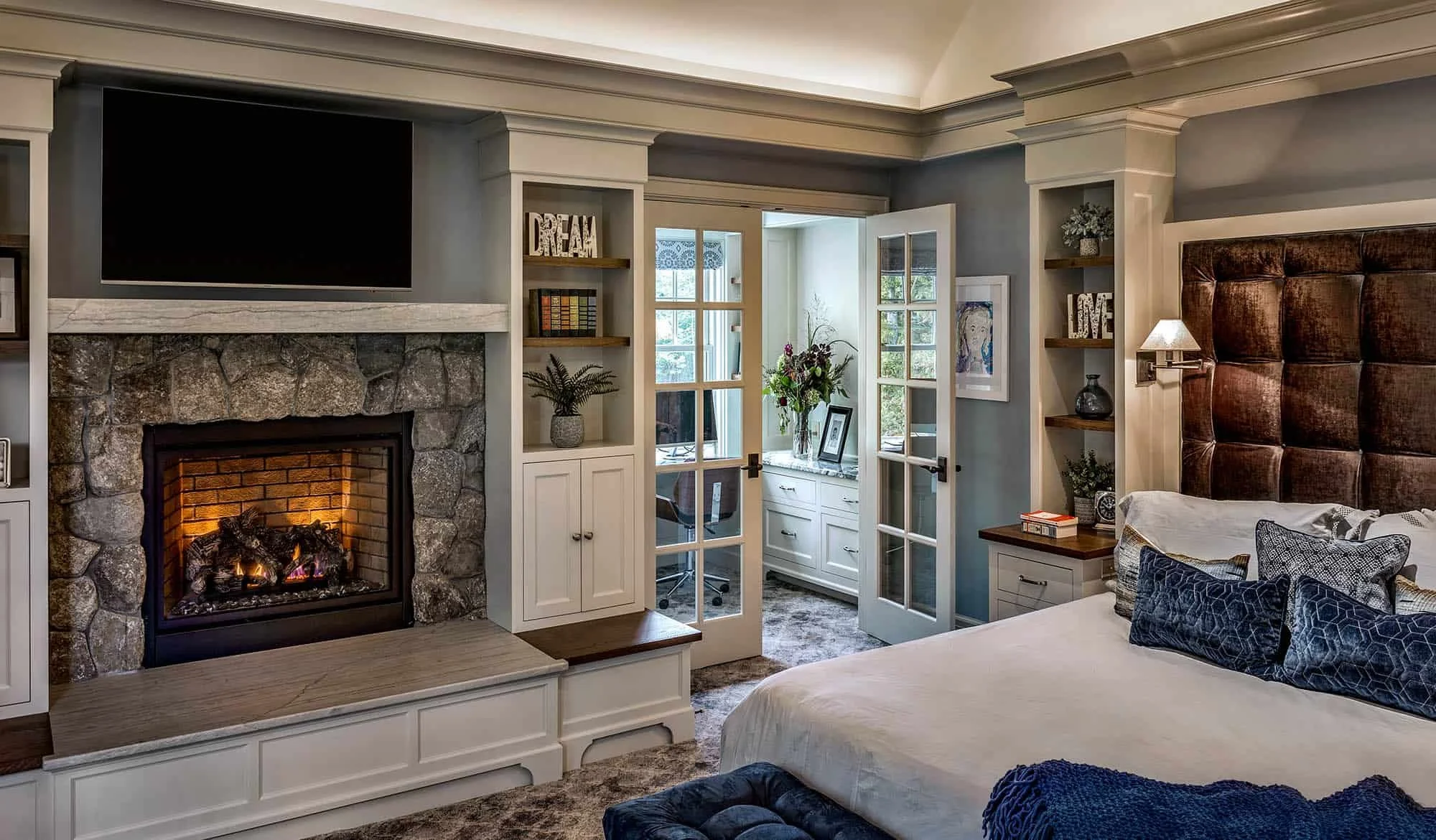 A cozy bedroom with a stone fireplace, a large dark brown headboard, bookshelves with decor, a glass door leading to a room with white furniture and a window with a view of greenery.