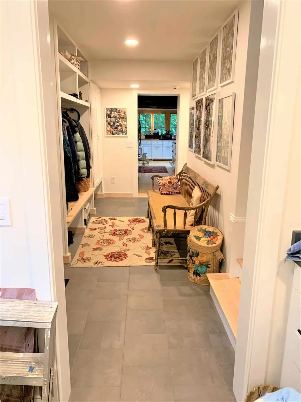 View of a small laundry or mudroom with a bench, coat hooks, and shelves for shoes and storage, leading to a kitchen area with large windows.