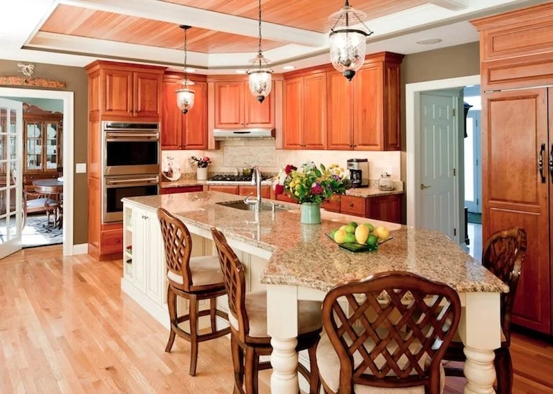 A spacious kitchen with wooden cabinets, a granite island, and pendant lighting. There is a stainless steel oven, a vase of flowers, and a bowl of lemons on the island.
