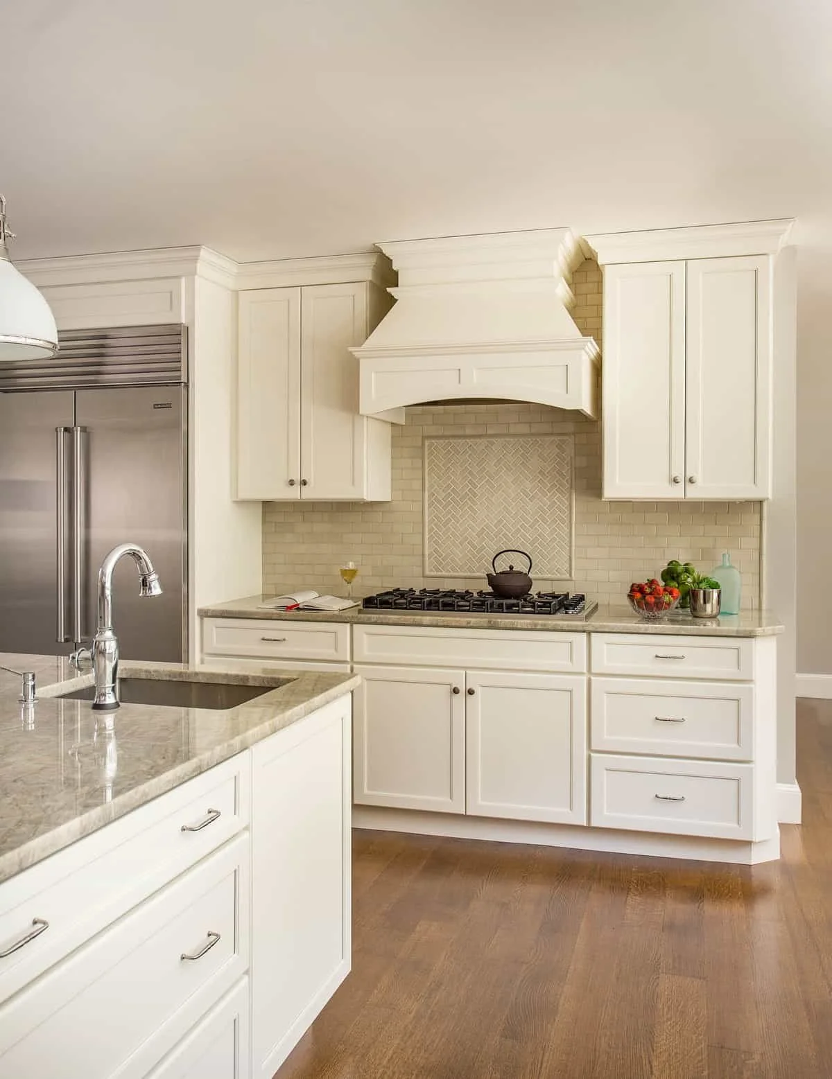 Kitchen with white cabinets, beige backsplash, gas stove, kitchen island with sink, and various kitchen items like a bowl of fruit, glass, book, and bottles.