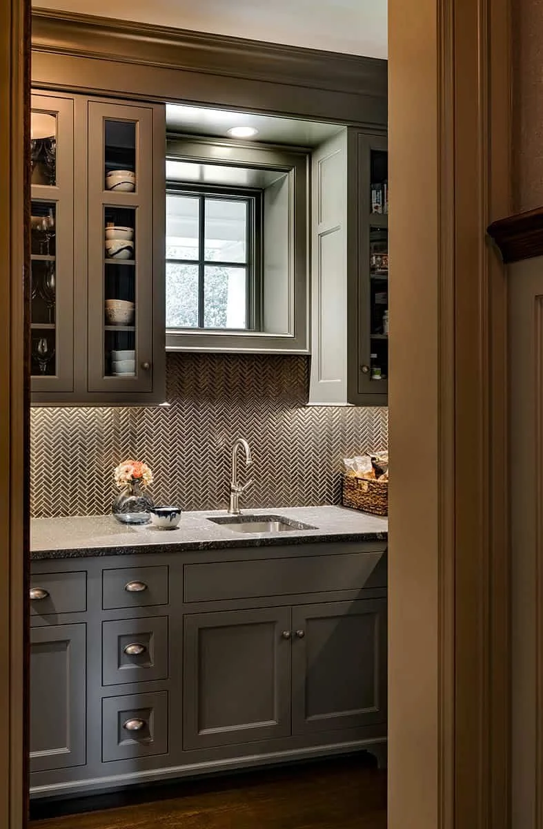 A small kitchen sink area with gray cabinets, a window above the sink, a herringbone-patterned backsplash, a small vase with flowers, a basket of snacks, and a small bowl.