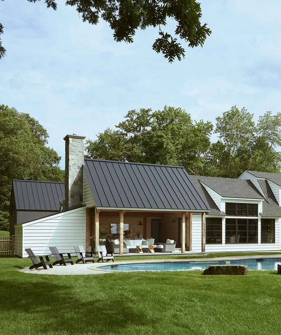 A backyard view of a modern house with a swimming pool, lawn chairs, and a patio with outdoor seating, trees in the background, and a partly cloudy sky above.