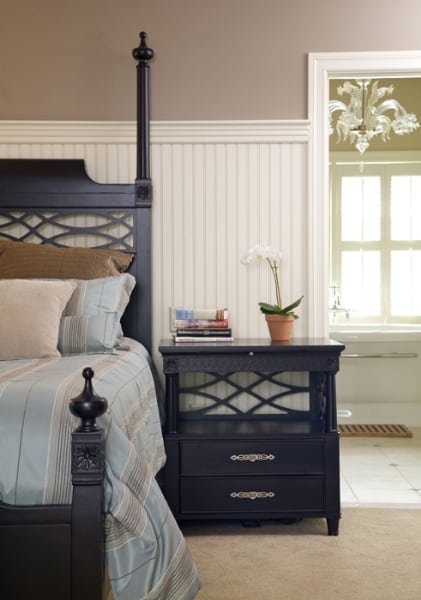 Bedroom with a black four-poster bed, beige bedsheets, and pillows; black nightstand with a stack of books and a white orchid plant; window with white trim and a view of an adjacent room.