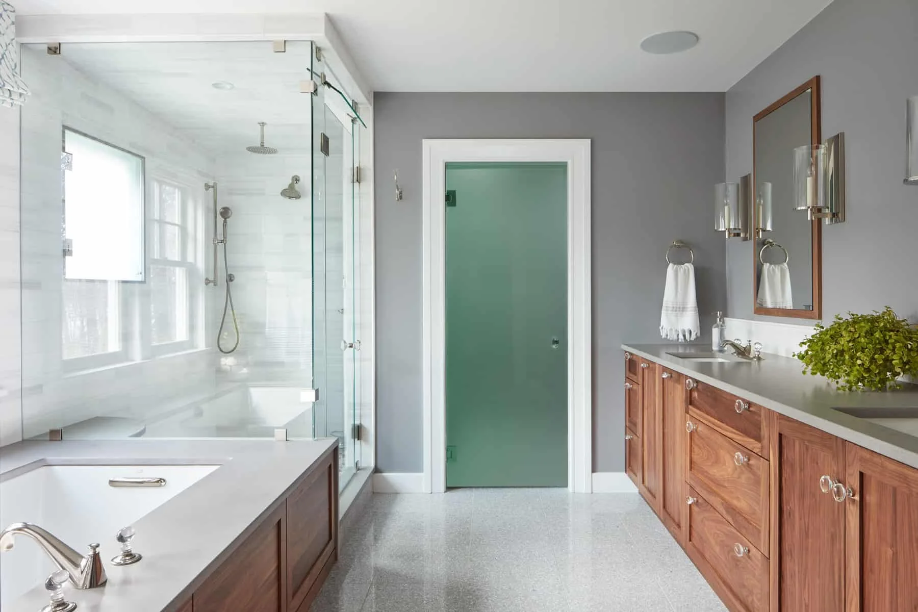 A bathroom with a glass shower enclosure, gray walls, wooden vanity with a white countertop, and a large mirror. There are two wall-mounted light fixtures, a towel hanging on a ring, a green door, and a potted plant on the countertop.