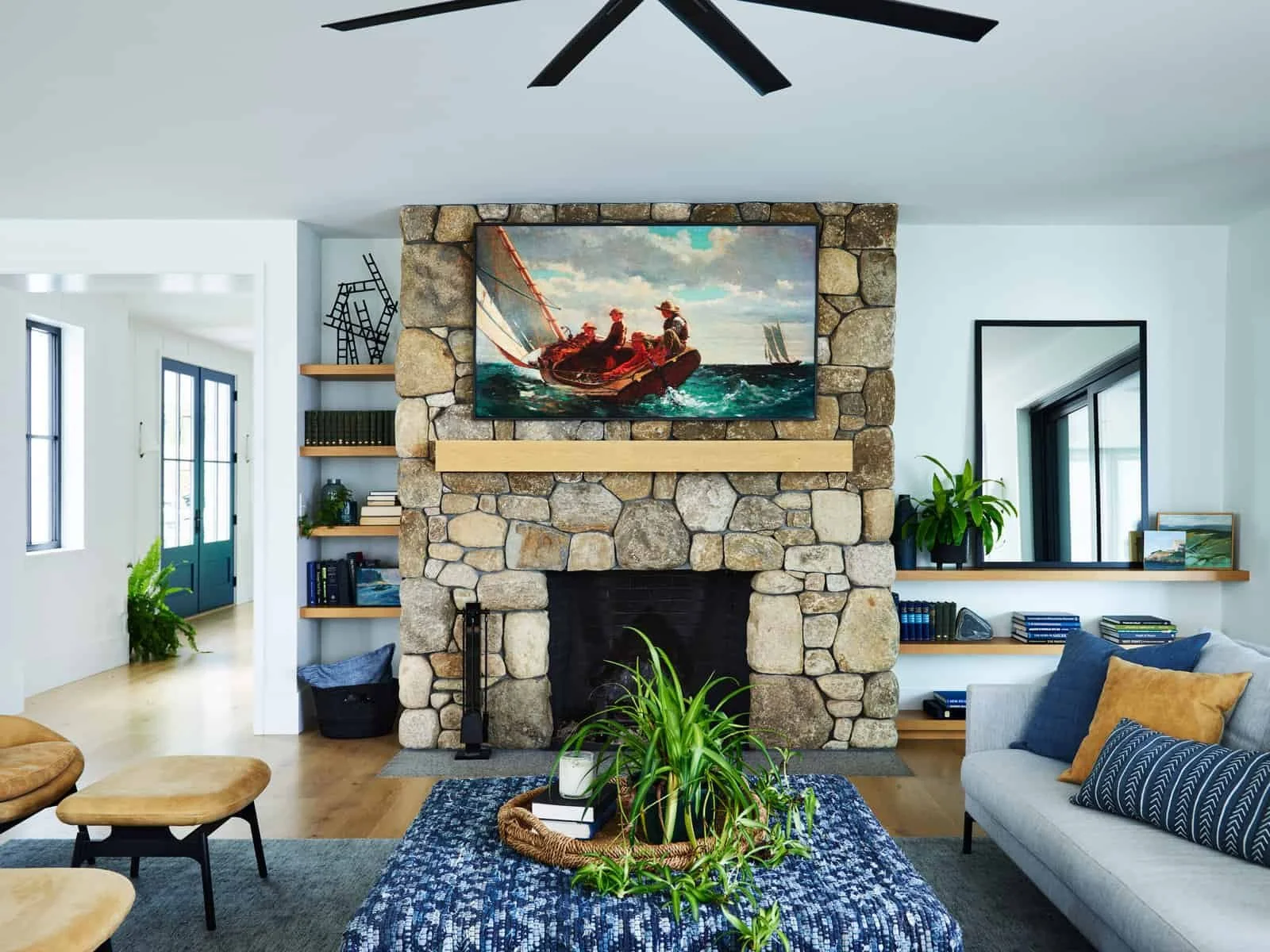 A living room with a stone fireplace, a painting of sailboats on the sea above the fireplace, and a large mirror on the right wall. There are plants, books, and furniture including a beige chair, a gray sofa, and a blue patterned rug.