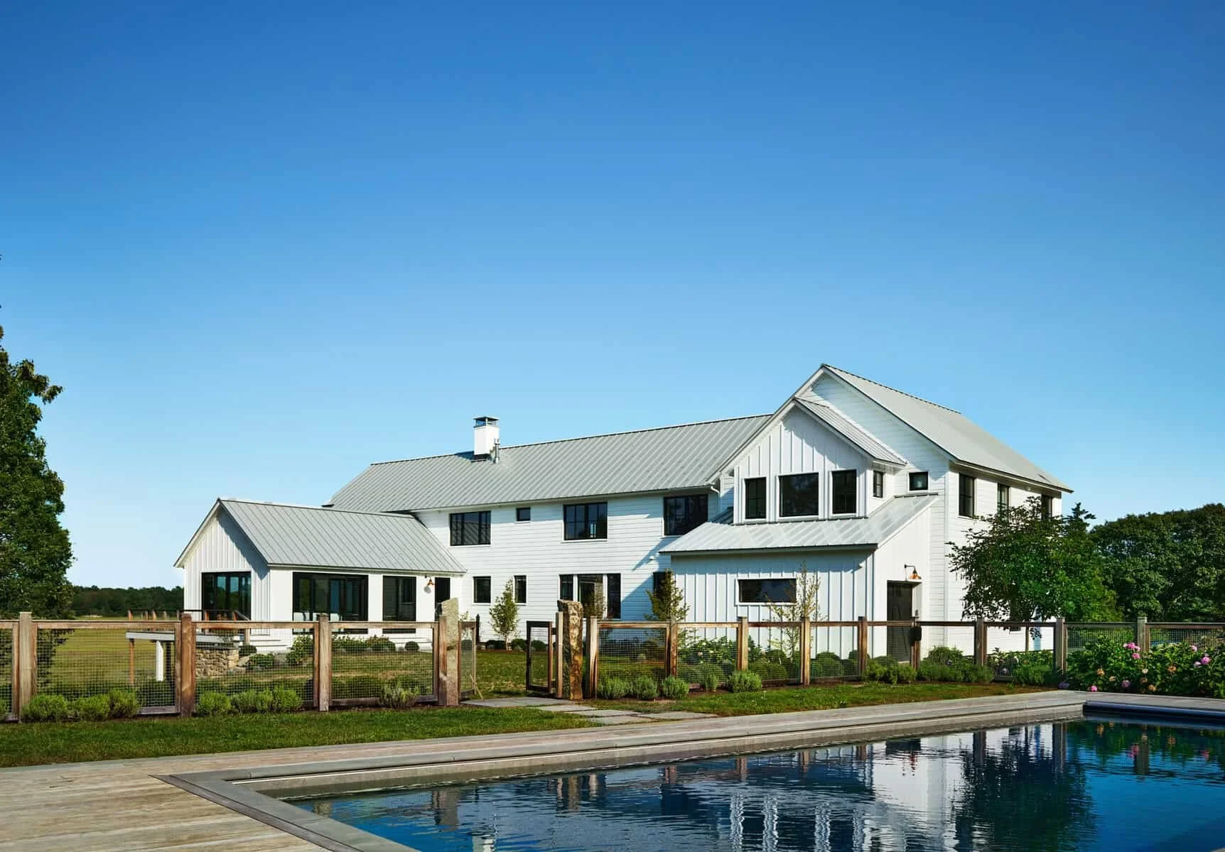 White modern house with black window frames and a gray metal roof, fenced yard, near a swimming pool, surrounded by greenery under a clear blue sky.