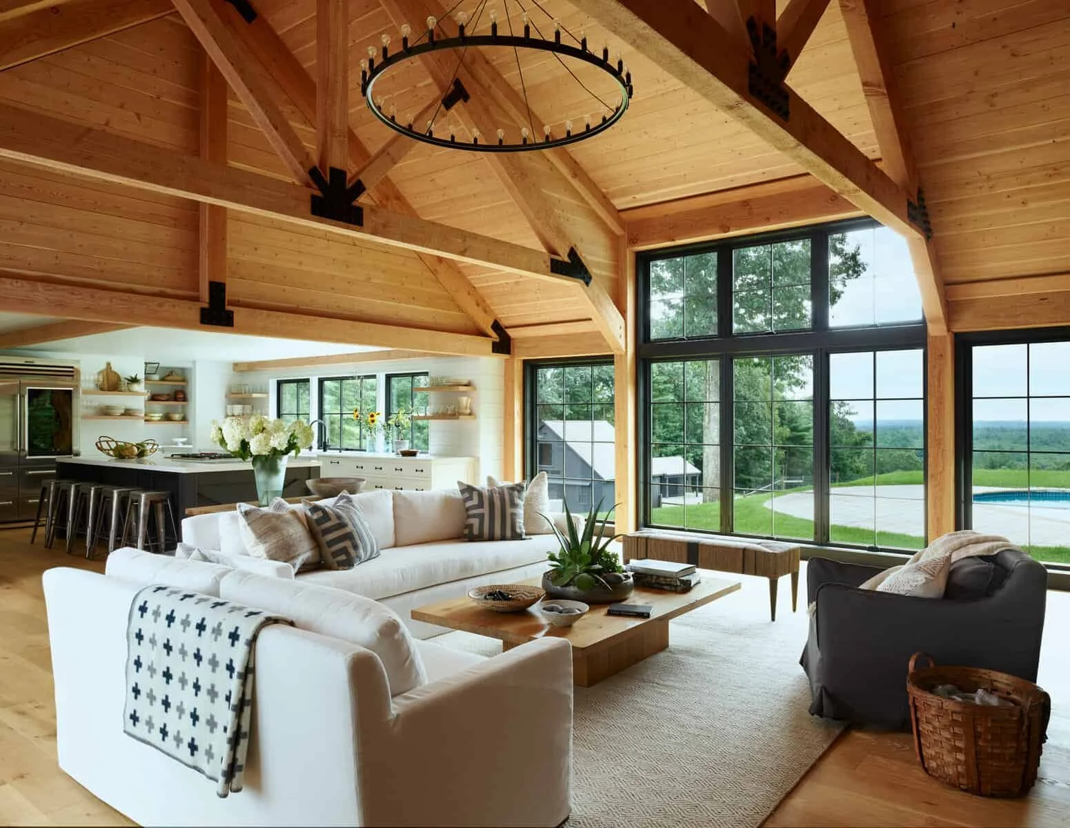 Interior of a spacious living room with large windows showing a backyard with a pool and trees. The room has a high wooden ceiling with exposed beams and a circular chandelier. There are white sofas with pillows, a wooden coffee table with decor, and