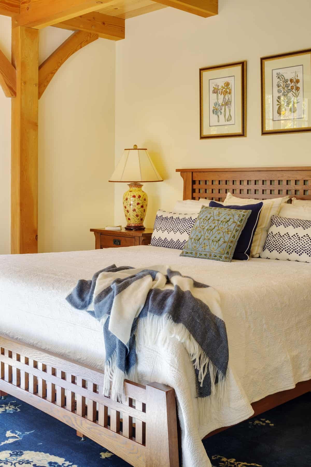 A neatly made bed with a wooden headboard, decorative pillows, a white blanket, and a striped throw blanket. A bedside table with a floral table lamp and framed botanical art on the wall in the background.
