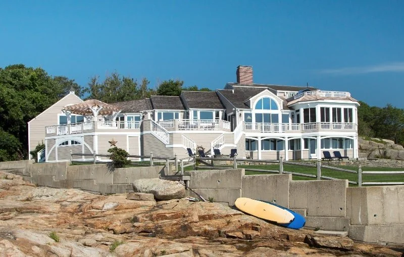 Large seaside house on elevated terrain with multiple decks, white railings, and a boat on rocky ground in the foreground.