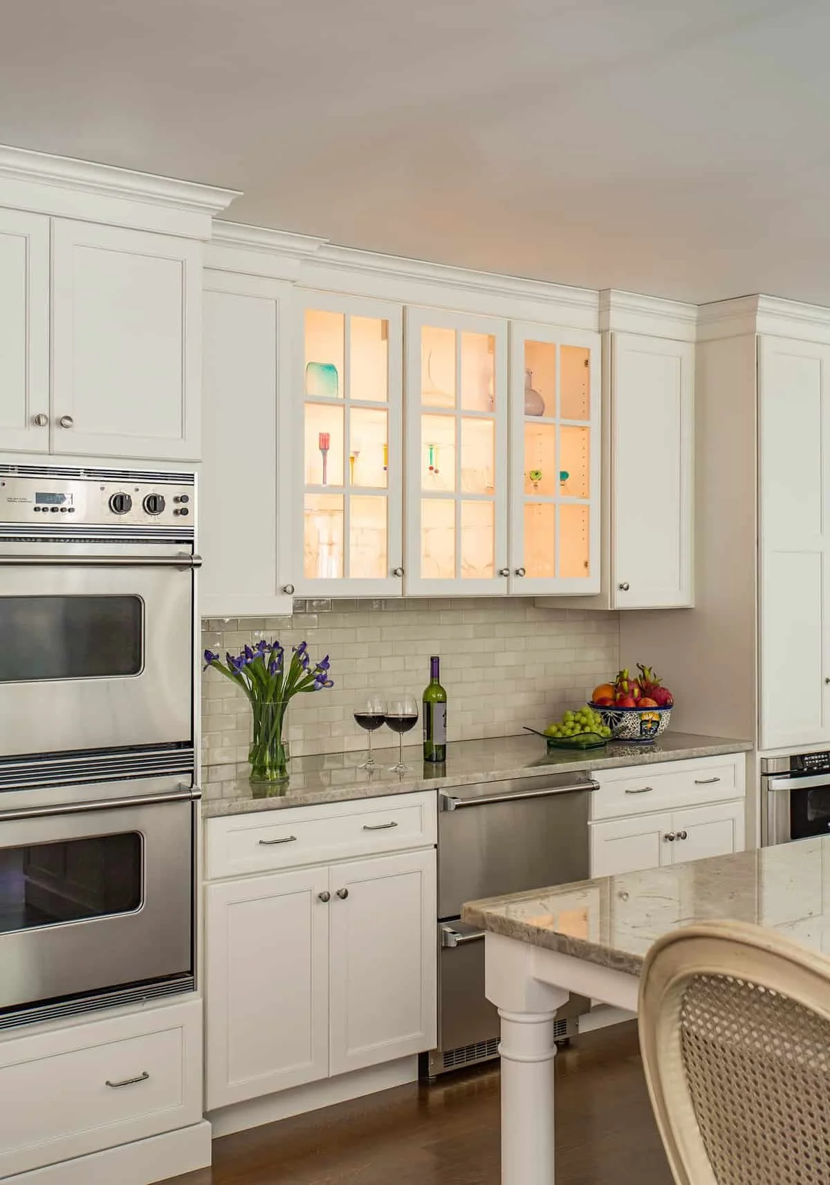 Kitchen with white cabinetry, stainless steel oven and microwave, glass-front cabinets, flower vase with purple flowers, two glasses of red wine, bottle of wine, bowl of grapes and apples on counter, beige countertop, beige tile backsplash, part of a