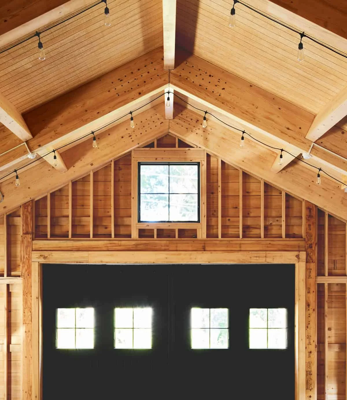 Interior of a wooden barn with a vaulted ceiling, string lights hanging, a window at the back, and a large garage door with small windows.