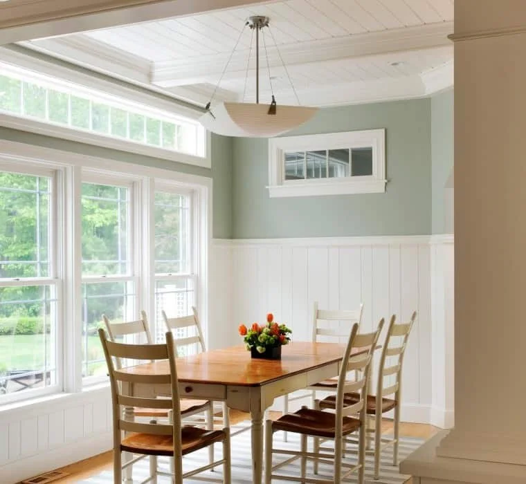 Dining room with a wooden table, six white chairs, a flower arrangement, large windows, and a light gray wall with white wainscoting.
