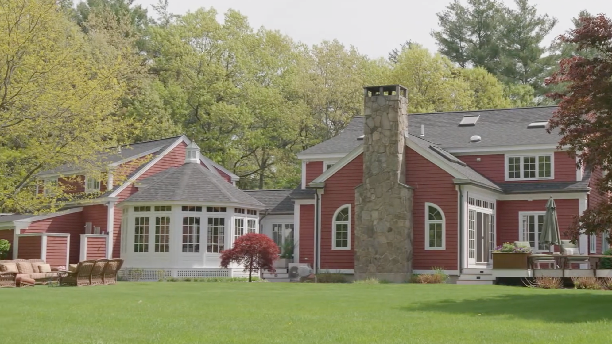 A large red traditional style house with white trim, a stone chimney, sunroom, and multiple roof skylights, surrounded by lush green trees and a well-maintained lawn.