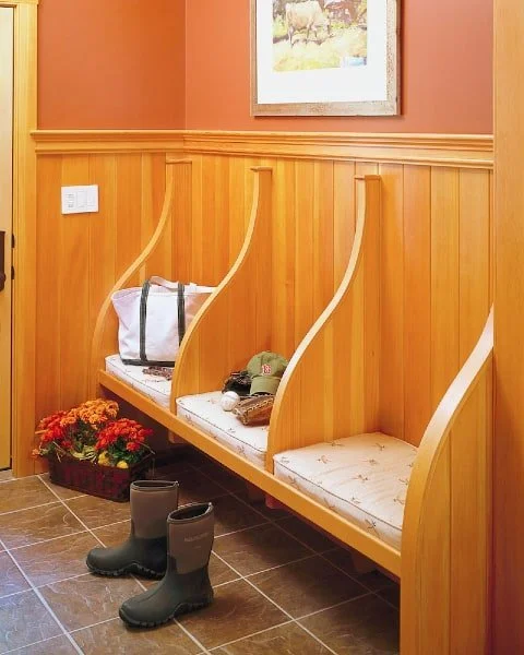 Wooden bench with three sections, each with a small cushion and separated by curved dividers, in a room with wood-paneled walls, a framed picture, and a pair of black rubber boots on the floor.