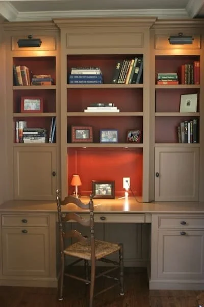 Custom built-in wooden bookshelf with closed cabinets at the bottom, open shelves in the middle, and picture frames and books displayed. A desk with drawers is in front, with a wooden chair. There are two small spotlights above the bookshelf.