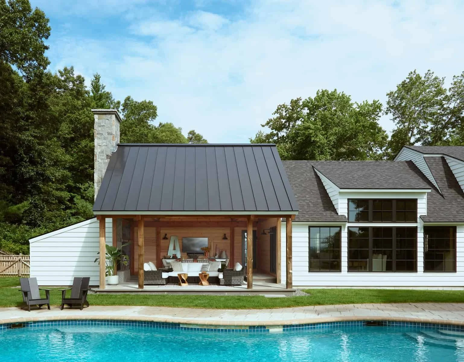 Backyard view of a house with a swimming pool in the foreground, a covered outdoor living area with furniture, and a house with large windows and a sloped roof in the background, surrounded by trees.