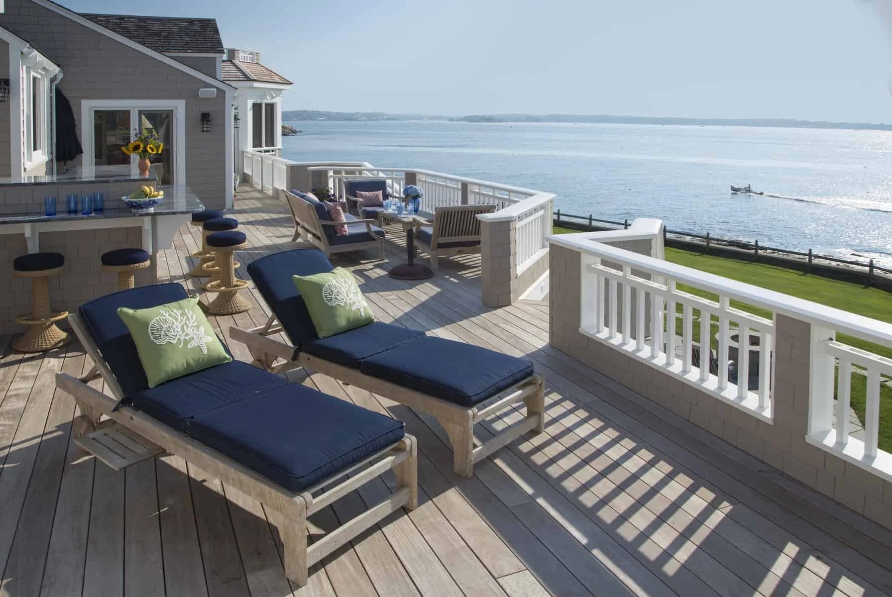 A spacious outdoor deck overlooking water with lounge chairs, a table with sunflowers, and a seating area with cushions and water views.