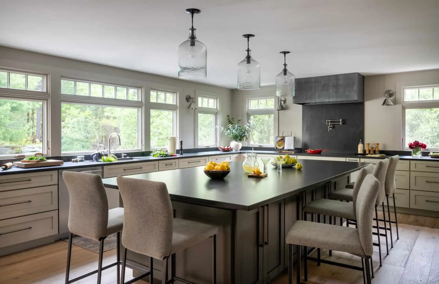 A modern kitchen with a large central island, light-colored cabinets, multiple windows, pendant lights, and various kitchenware and decorative items on the counters.