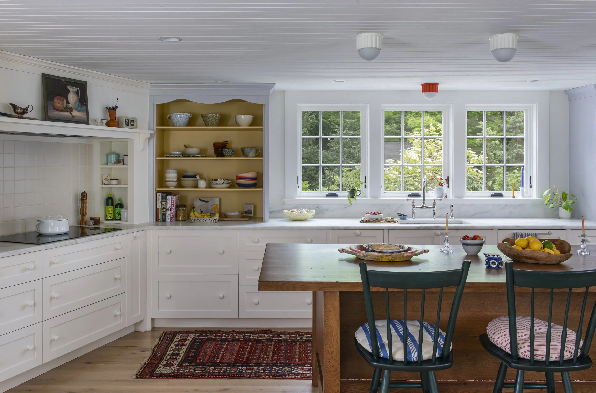 A bright kitchen with windows overlooking green trees, white cabinetry, open shelves with bowls and decor, a marble countertop, a wooden table with a bowl of lemons, and a colorful rug.