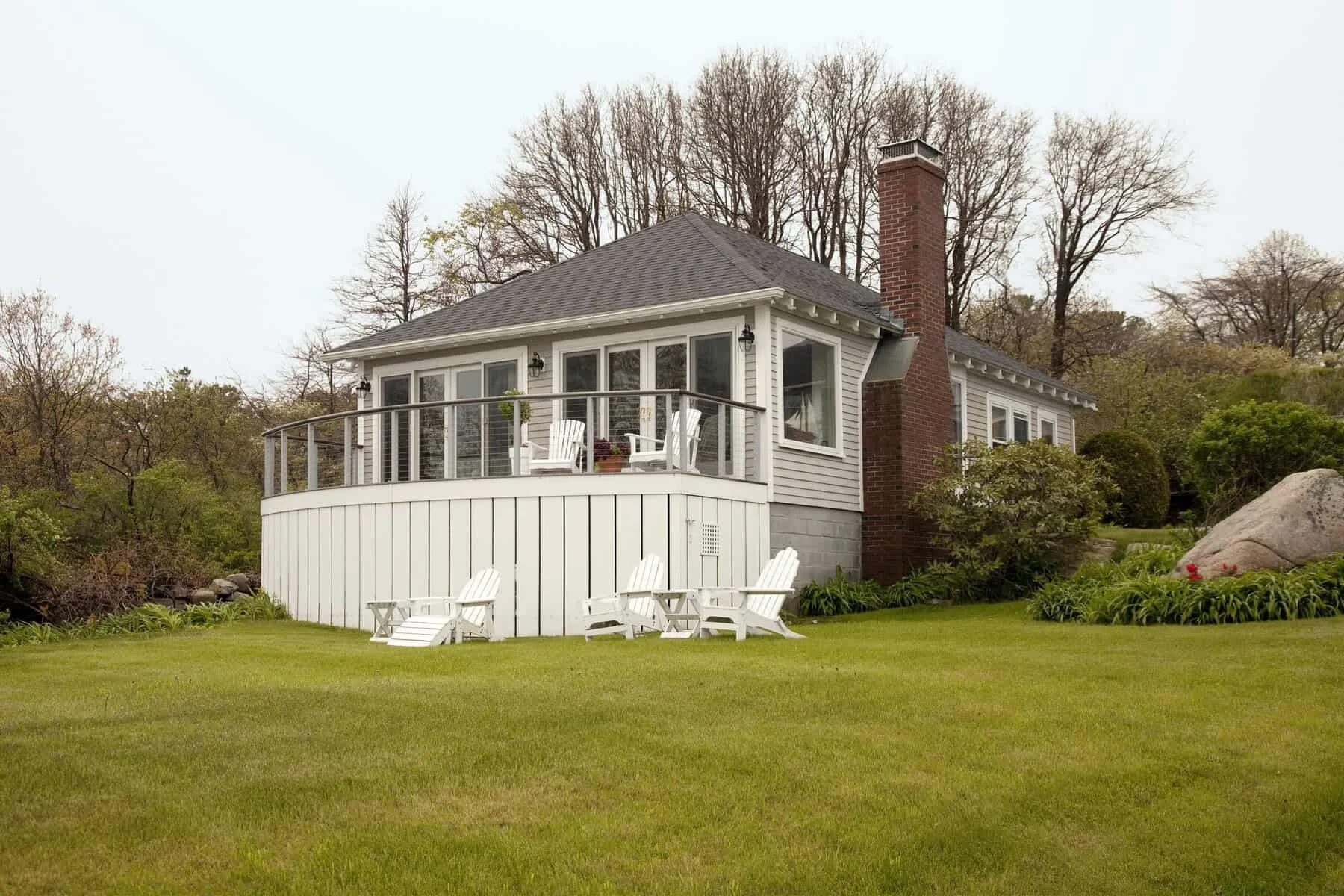 A house with a deck, white chairs, and a lush green lawn, surrounded by trees and landscaping.