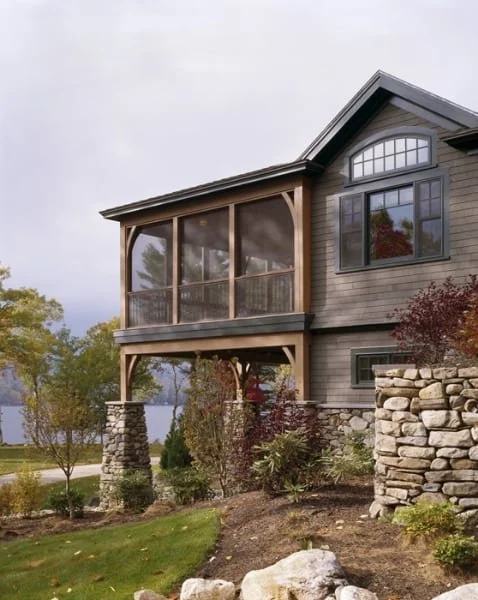 A two-story house with a screened porch on the upper level, gray wooden siding, large windows, and stone pillars supporting the porch, surrounded by landscaped yard with trees and rocks.
